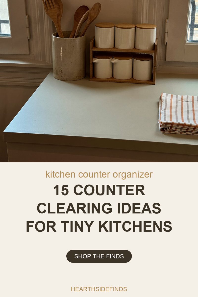 Photo of a bare cream kitchen counter corner in daylight from two windows, one at each side, with a panelled painted wall and window frames behind. At the back left a tall straight-sided speckled stoneware crock holds four wooden utensils handles down and heads up: a fork and three spoons. Beside it a two-tier wooden riser holds six cream ceramic canisters with round wooden lids, three on the top tier and three on the lower one. A white tea towel with orange and grey stripes lies folded at the right edge. The rest of the counter is empty.