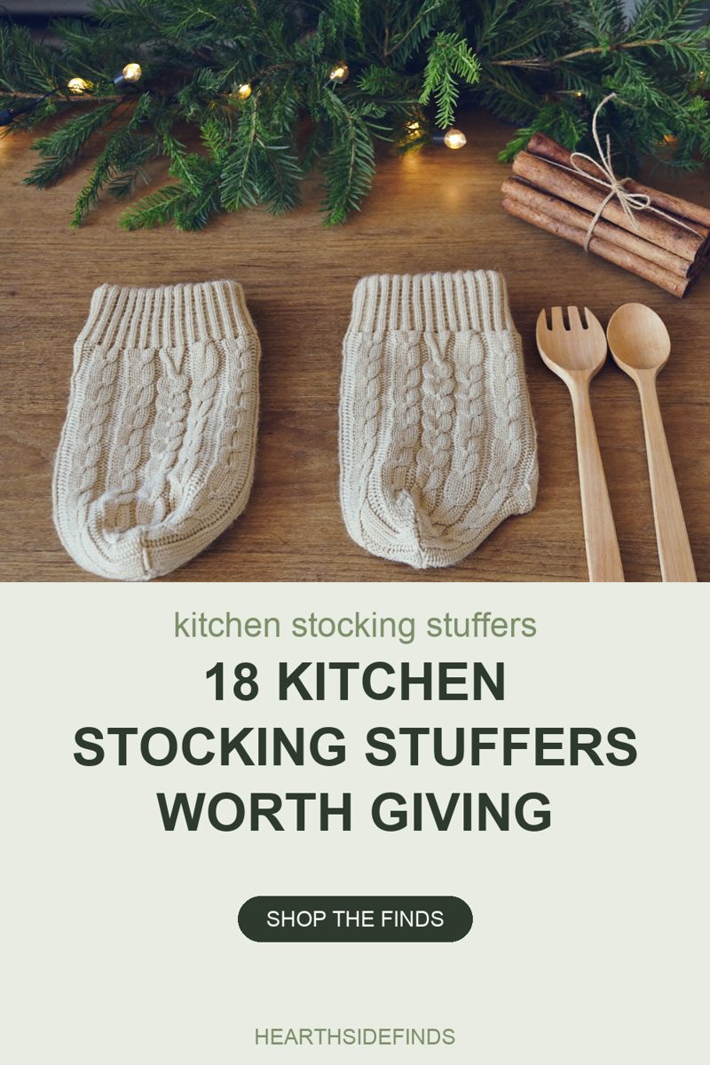 Overhead photo on an oak table of two cream cable-knit pouches with ribbed cuffs and rounded closed ends lying flat side by side, a wooden salad fork and serving spoon laid beside them on the right, a bundle of cinnamon sticks tied with twine in the top right corner, and fir branches strung with small warm fairy lights across the top of the frame