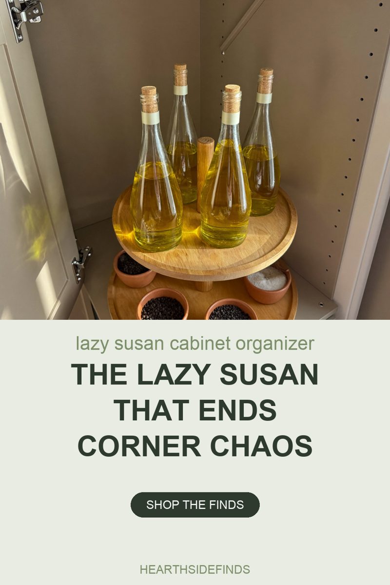Photo into an open cream corner cabinet where a two-tier wooden turntable holds four tall clear bottles of golden oil with cork stoppers on the upper tier, and small terracotta pinch bowls of peppercorns and coarse salt on the lower tier, lit by daylight from the left